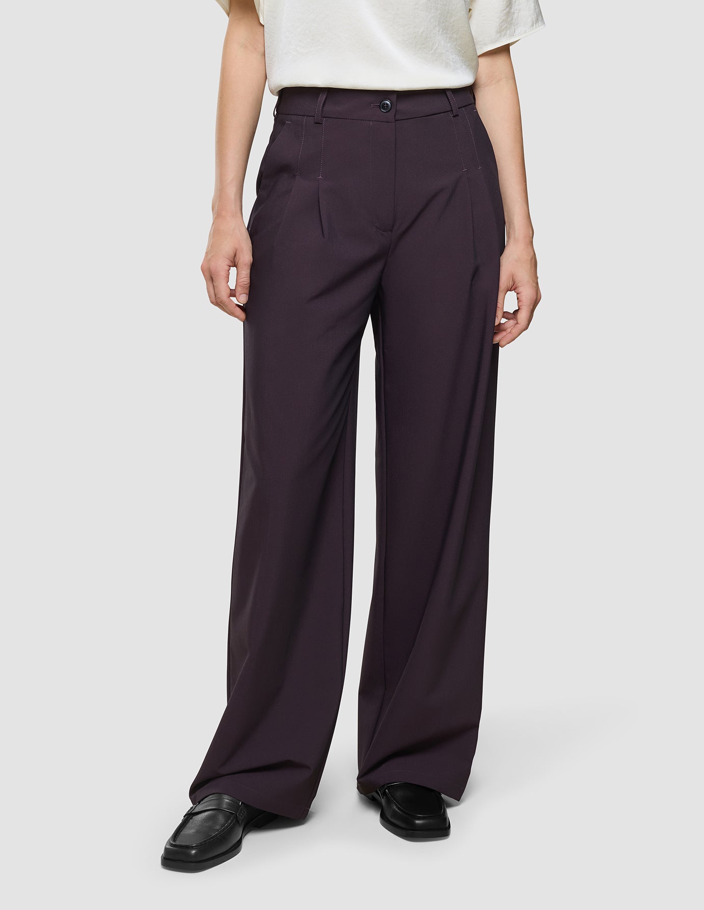 En person iført en hvid T-shirt, der er stoppet ned i et par Avira-bukser til kvinder i modellen »Wide« i farven »Burgundy Berry«, samt sorte loafers.