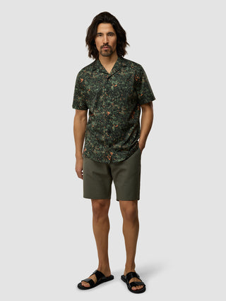 En mand iført en kortærmet Bowling-skjorte i farven »Forest Night«, mørke shorts og sorte sandaler.