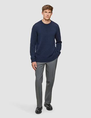En mand har en tyk, blød uldstrikket sweater med rund hals i mørk marineblå melange på sammen med grå dressbukser og sorte sko.