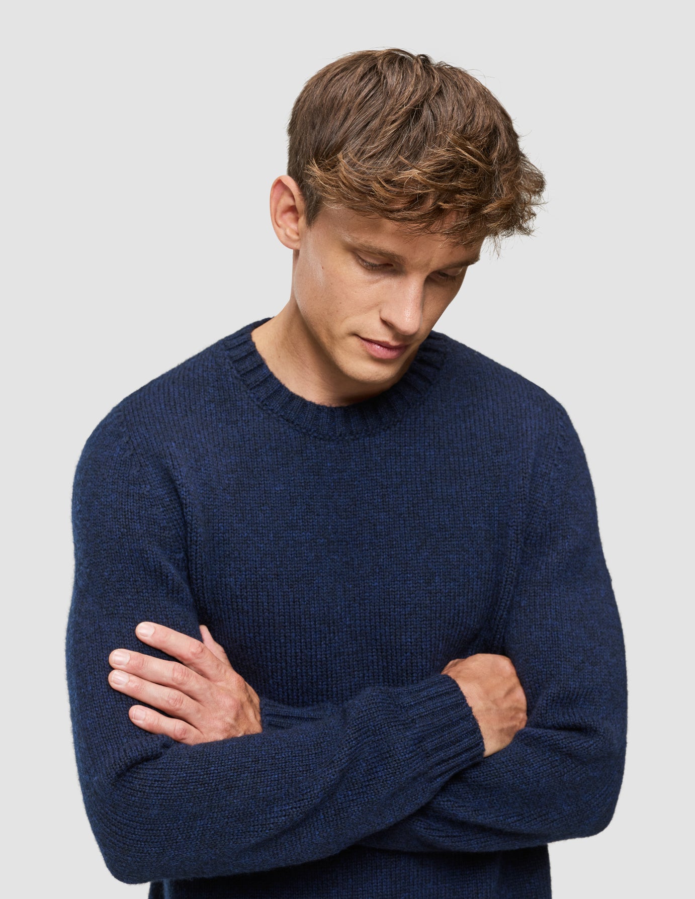 En mand med armene over kors, iført en tyk, blød uldstrikket sweater med rund hals i mørk marineblå melange.