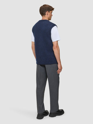 En mand set bagfra, iført en tyk, blød uldstrikket vest i mørk marineblå melange over en hvid T-shirt, grå bukser og sko.