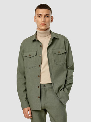 En mand har en beige rullekravebluse på under en Essential Overshirt i farven Nightfall Green Melange og et par bukser i samme farve.