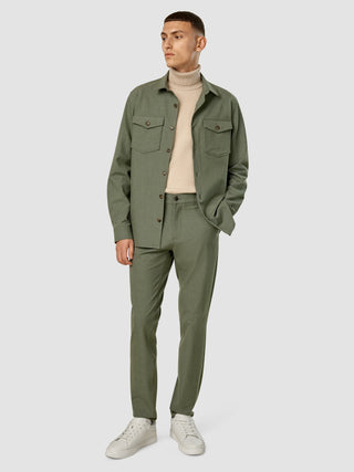 En mand iført en Essential Overshirt i farven Nightfall Green Melange, en beige rullekravebluse, matchende bukser og hvide sneakers.