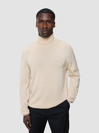 En mand iført en Seamless Premium Merino-strikket rullekravebluse i Birchwood og sorte bukser.