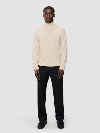 En mand iført en Seamless Premium Merino-strikket rullekravebluse i farven Birchwood, sorte bukser og sorte sneakers.