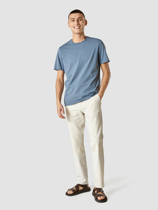 En smilende mand iført en Supima-T-shirt i farven »Blue Mirage«, hvide bukser og mørke sandaler.
