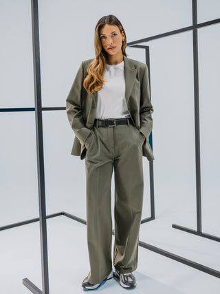 En kvinde iført en salviegrøn Essential Blazer i farven Dark Olive og en hvid T-shirt, der står midt blandt sorte metalrammer.