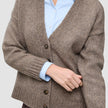 En kvinde har en tyk, blød uldstrikket cardigan i farven taupe-melange på over en lyseblå skjorte med krave.