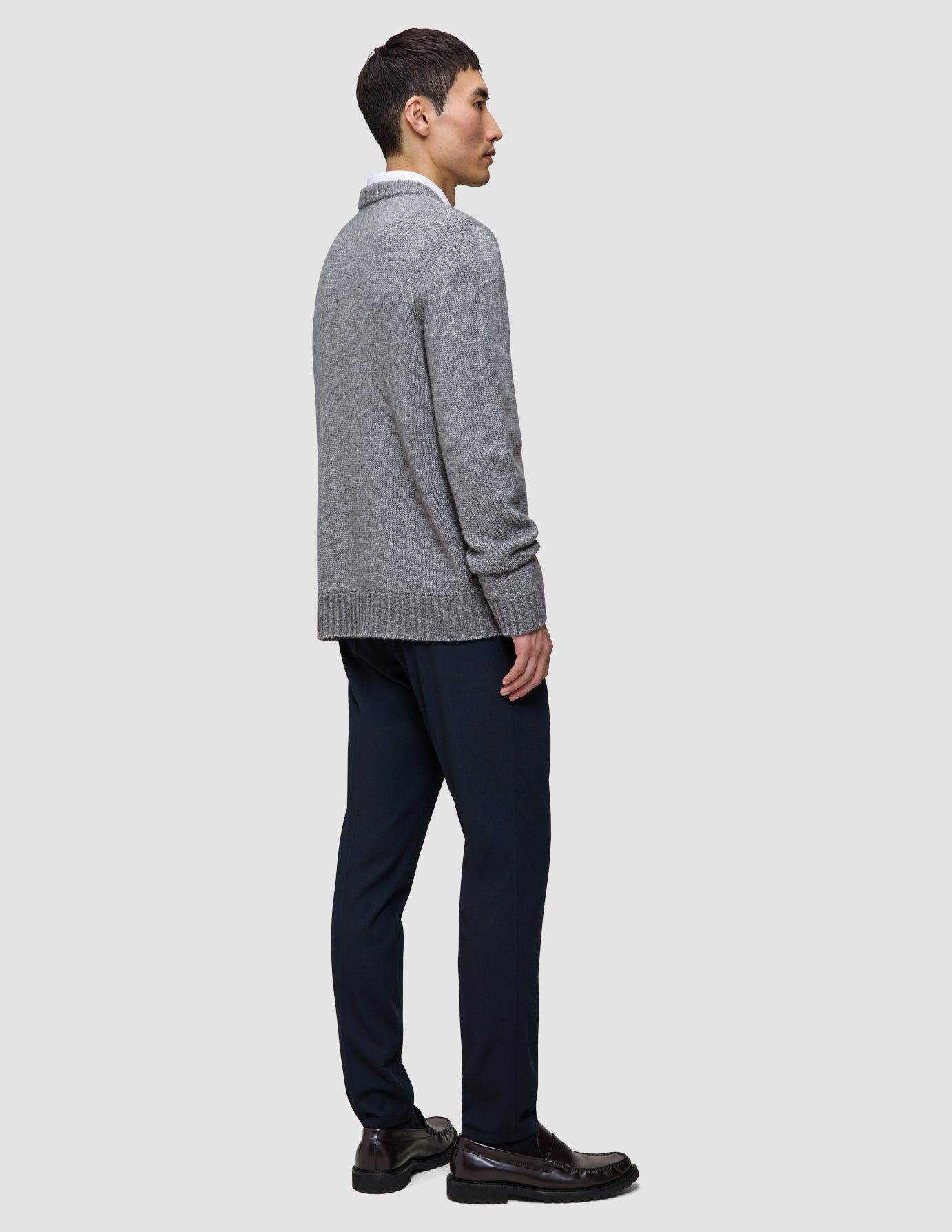 Set fra siden: En person iført en grå, tyk, blød uldstrikket sweater med rund hals i farven Glacier Grey, mørke bukser og mørke loafers.