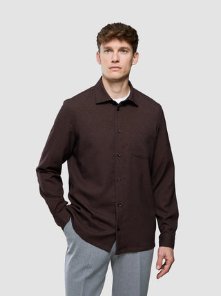 En mand iført en mørkebrun Effortless-skjorte Dark Brown over en hvid undertrøje og grå bukser.