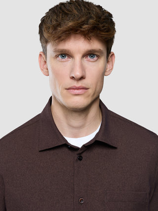En mand iført en mørkebrun »Effortless Shirt« og en hvid undertrøje, der ser direkte ind i kameraet.