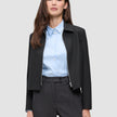 En person, der har en sort Essential Short Zip Jacket på over en lyseblå skjorte med knapper og grå bukser.