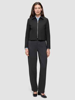 En kvinde iført en sort Essential Short Zip Jacket over en lyseblå skjorte med krave og mørke bukser.