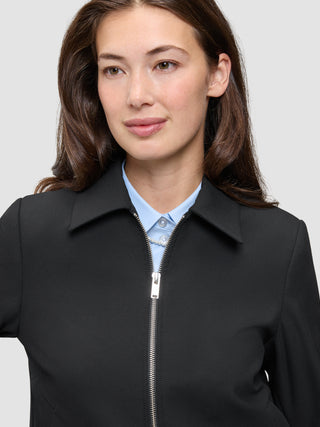 En person, der har en sort Essential Short Zip Jacket på over en lyseblå skjorte med krave.