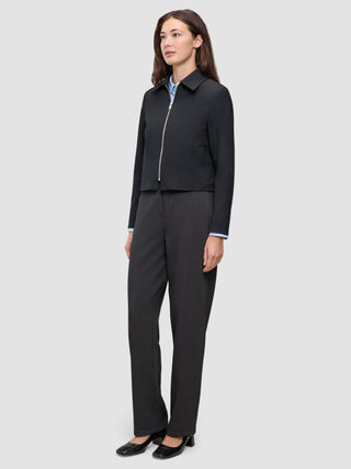 En kvinde iført en sort Essential Short Zip Jacket over en lyseblå skjorte med krave og mørkegrå bukser.