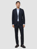 En mand iført Motion Suit Pants Regular Midnight Blue i et mørkeblåt jakkesæt, en lyseblå skjorte og mørke loafers.