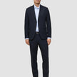 En mand iført Motion Suit Pants Regular Midnight Blue i et mørkeblåt jakkesæt, en lyseblå skjorte og mørke loafers.
