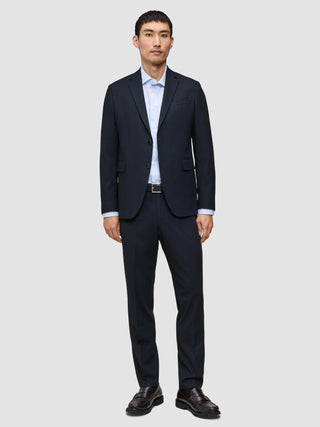 En mand iført Motion Suit Pants Regular Midnight Blue i et mørkeblåt jakkesæt, en lyseblå skjorte og mørke loafers.