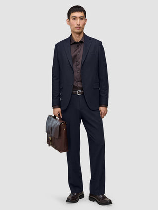En mand iført et par marineblå Motion Suit-bukser i afslappet pasform Midnight Blue, en mørk skjorte og loafers holder en brun lædermappe.