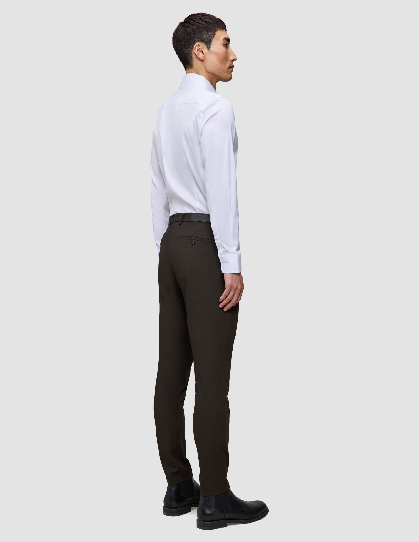 En mand set fra siden, iført en hvid skjorte med knapper og et par Motion Suit-bukser i Slim-pasform i farven Cognac med dansk design.