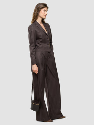 En kvinde set i profil er iført en »Women Raina Short Blazer« i farven Chestnut med nålestriber og et par matchende bukser, mens hun holder en lille taske.