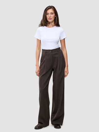 En person iført en hvid T-shirt og et par Serene Pants Wide Walnut Herringbone, som er mørkebrune bukser med brede ben og herringbone-mønster.