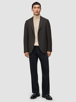 En mand i et look med strik og blazer bestående af en merino-trøje med rund hals, en mørk uldblazer med mønster, sorte bukser og sorte loafers.