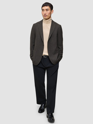 En person i et look med strik og blazer, iført en beige Premium Merino-trøje med rund hals, en mørk mønstret blazer og sorte loafers.