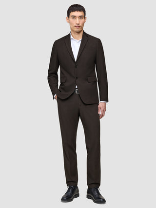 En mand er klædt i The suit that does it all i mørkebrun, en hvid skjorte med høj krave og sorte lædersko.