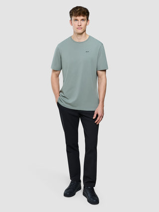 En mand iført en Supima Legacy T-shirt i farven »Mist Green«, sorte bukser og mørke sneakers.