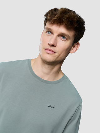 En mand iført en Supima Legacy-T-shirt i farven »Mist Green« med bogstaverne »Snt.« broderet på brystet.