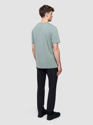 En mand set bagfra iført en Supima Legacy-T-shirt i farven »Mist Green«, sorte bukser og sorte sneakers.