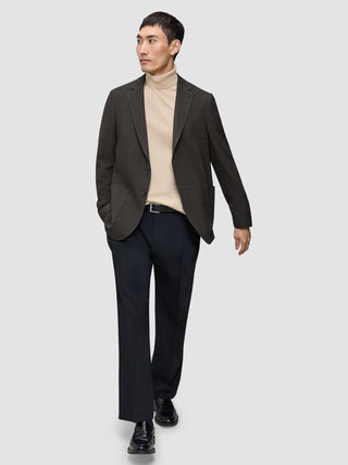 En mand har en mørkebrun, slim-fit Timeless-uldsblazer med tern på over en beige rullekravebluse og mørke bukser.