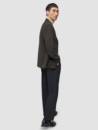 En mand står med siden til i en Timeless-uldsblazer i slim-fit med mørkebrunt ternet mønster, over en beige rullekravebluse og mørke bukser.