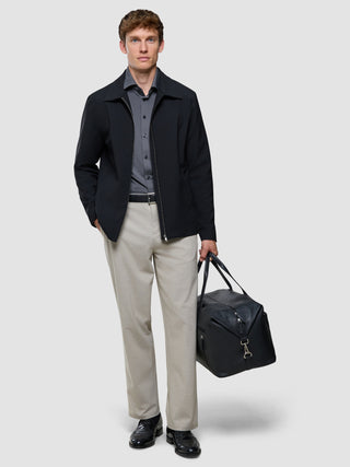 En mand i en sort jakke, en »Travel Shirt Charcoal Grey Slim« og lyse bukser står med en sort rejsetaske i hånden.