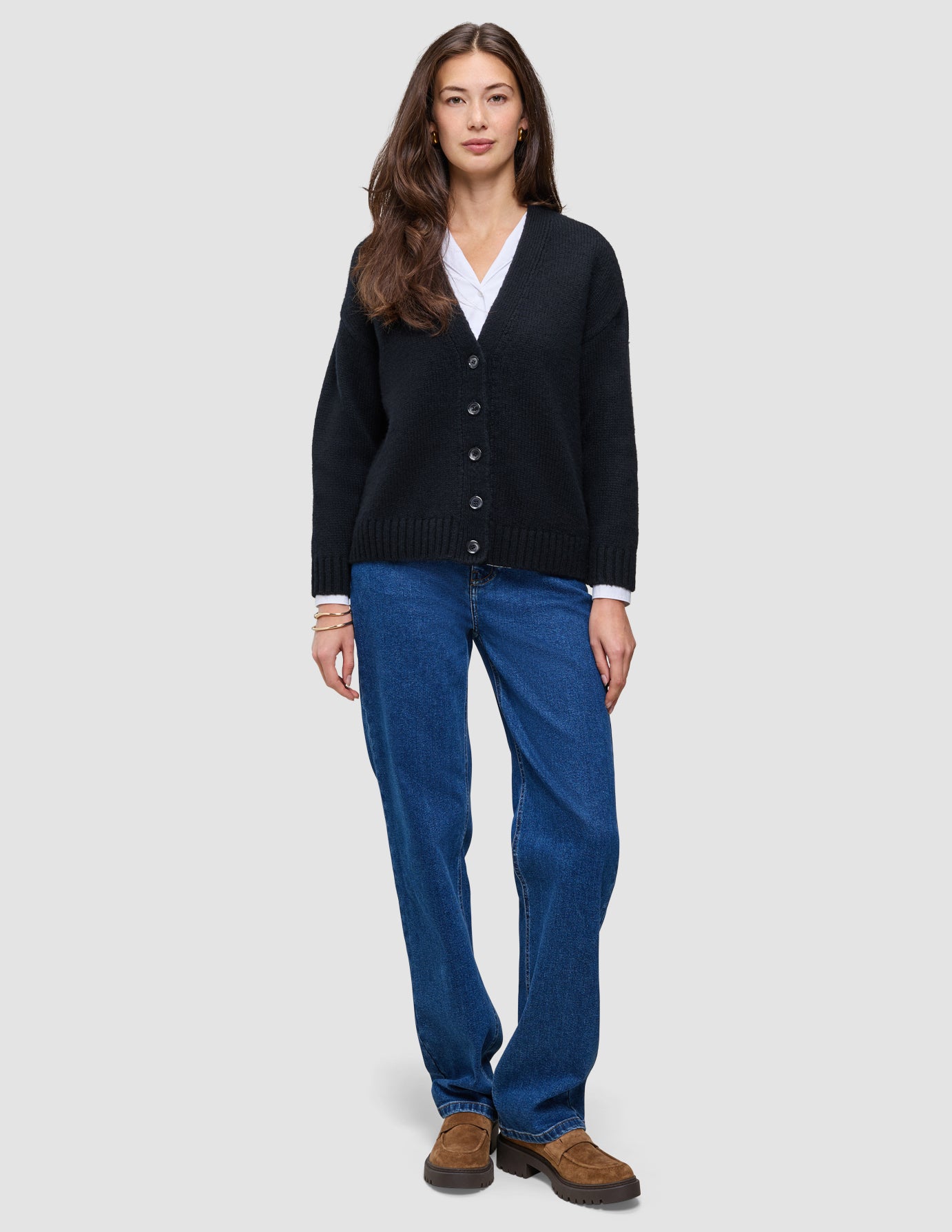 En kvinde iført en sort, tyk, blød uldstrikket cardigan fra serien Women over en hvid skjorte med krave og blå jeans.