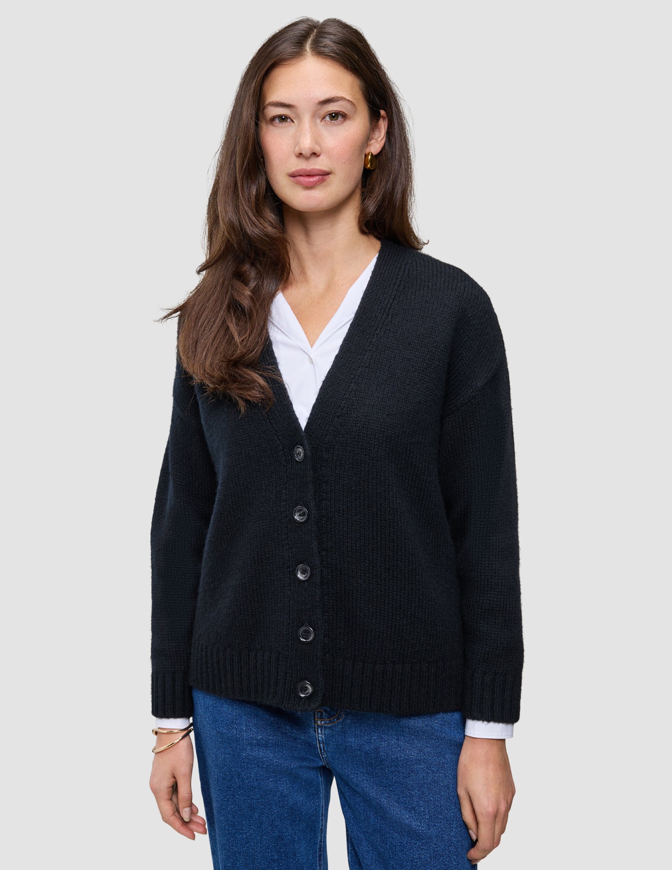 En kvinde iført en sort, tyk, blød uldstrikket cardigan fra serien Women over en hvid skjorte med krave og blå jeans.