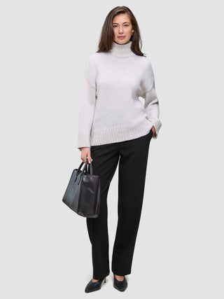 En kvinde i en beige rullekravesweater og et par sorte Women GO2-bukser med høj talje i lige snit, der holder en sort håndtaske.