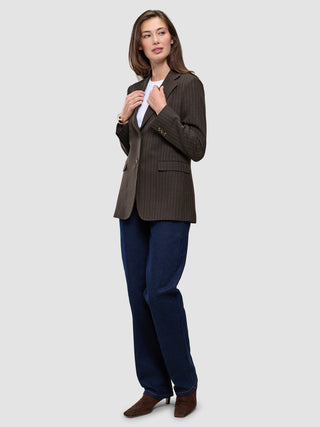 En kvinde har en brun Women Serene Blazer Regular Walnut Herringbone på over en hvid T-shirt, blå jeans og brune støvler.