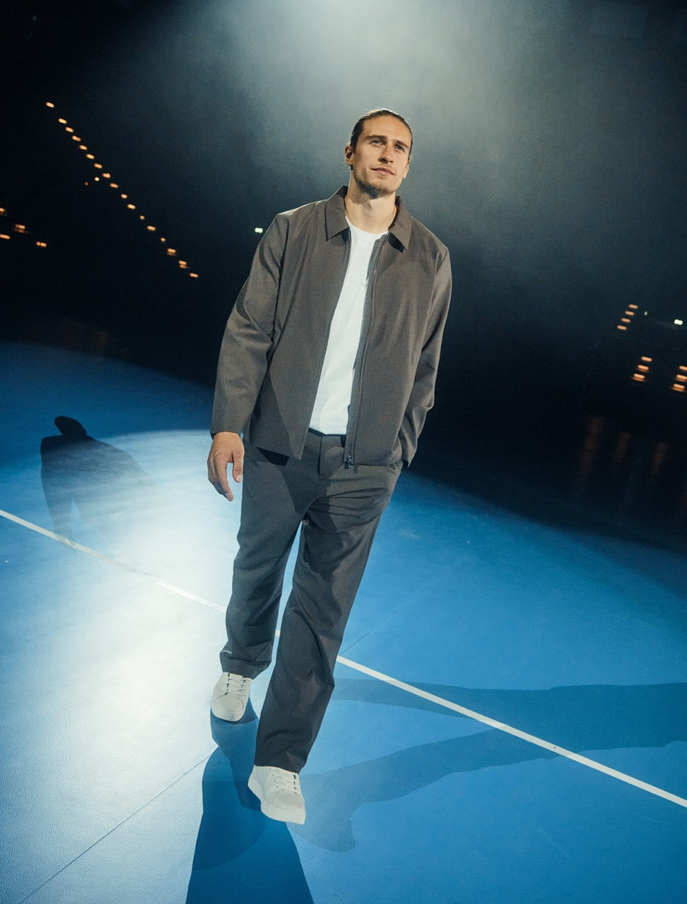 Mand i grå skjorte og hvide sneakers på blåt sportsgulv under spotlights.