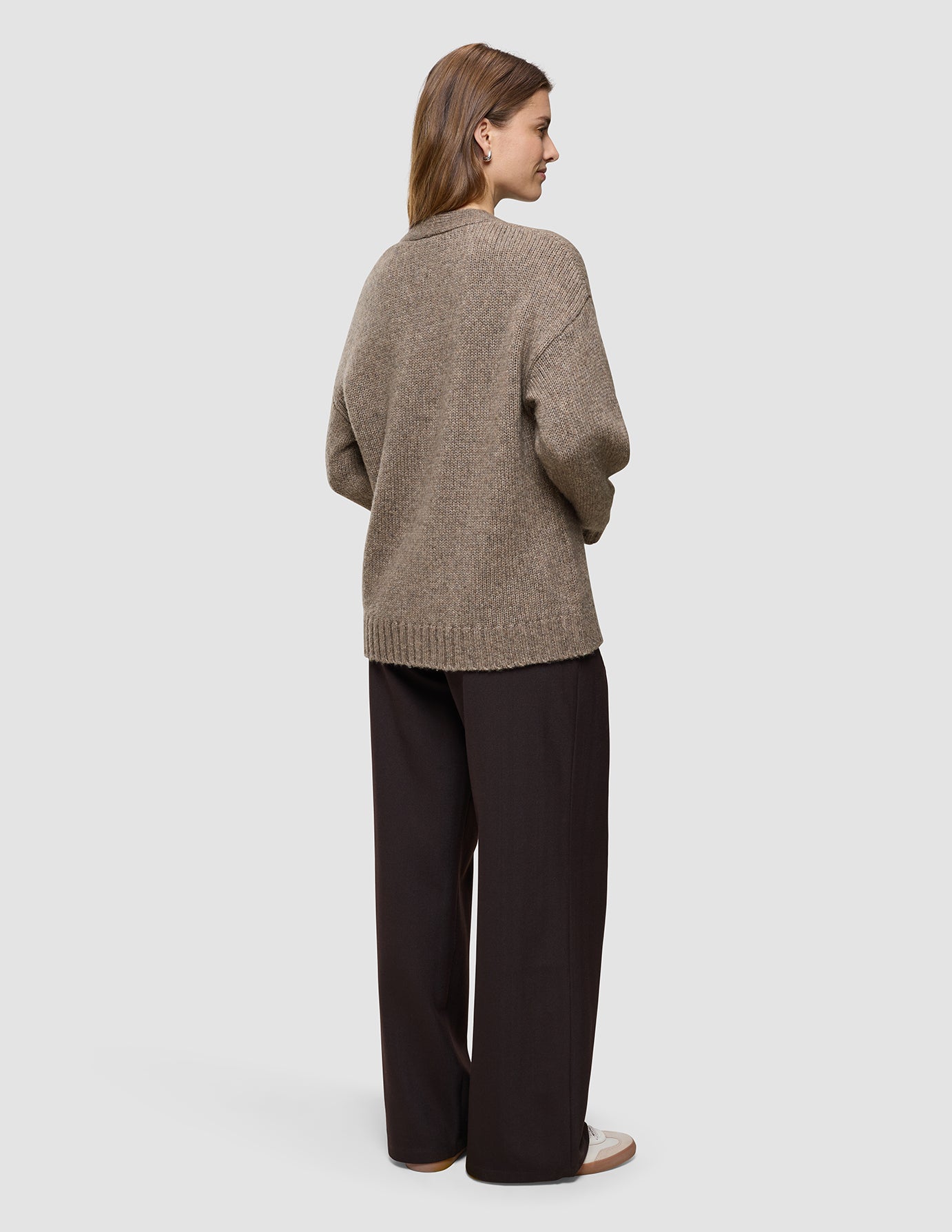 En person set bagfra, iført en tyk, blød uldstrikket cardigan til kvinder i farven taupe melange og mørkebrune bukser med brede ben.