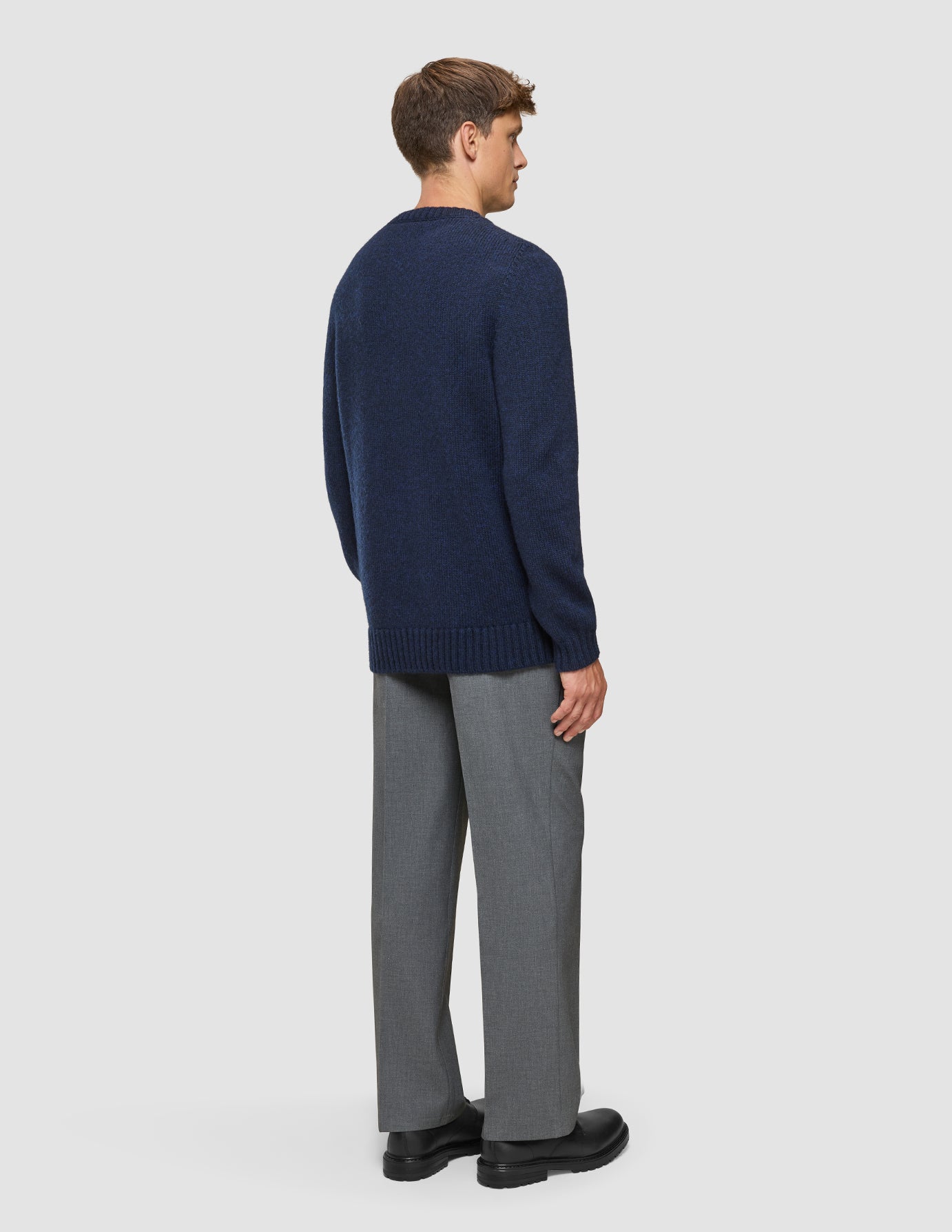 En mand set bagfra, iført en tyk, blød uldstrikket sweater med rund hals i mørk marineblå melange og grå bukser.