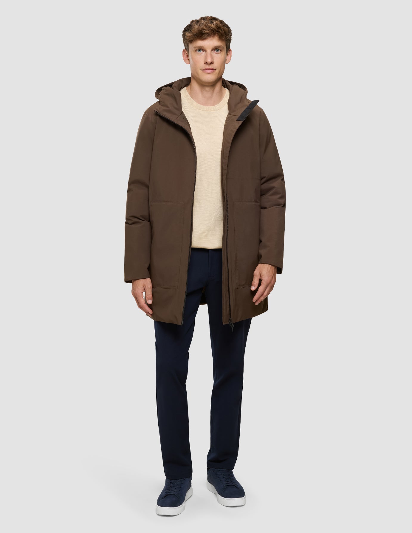 En mand iført en Utility Jacket i sadelbrun, en beige sweater, marineblå bukser og marineblå sneakers.