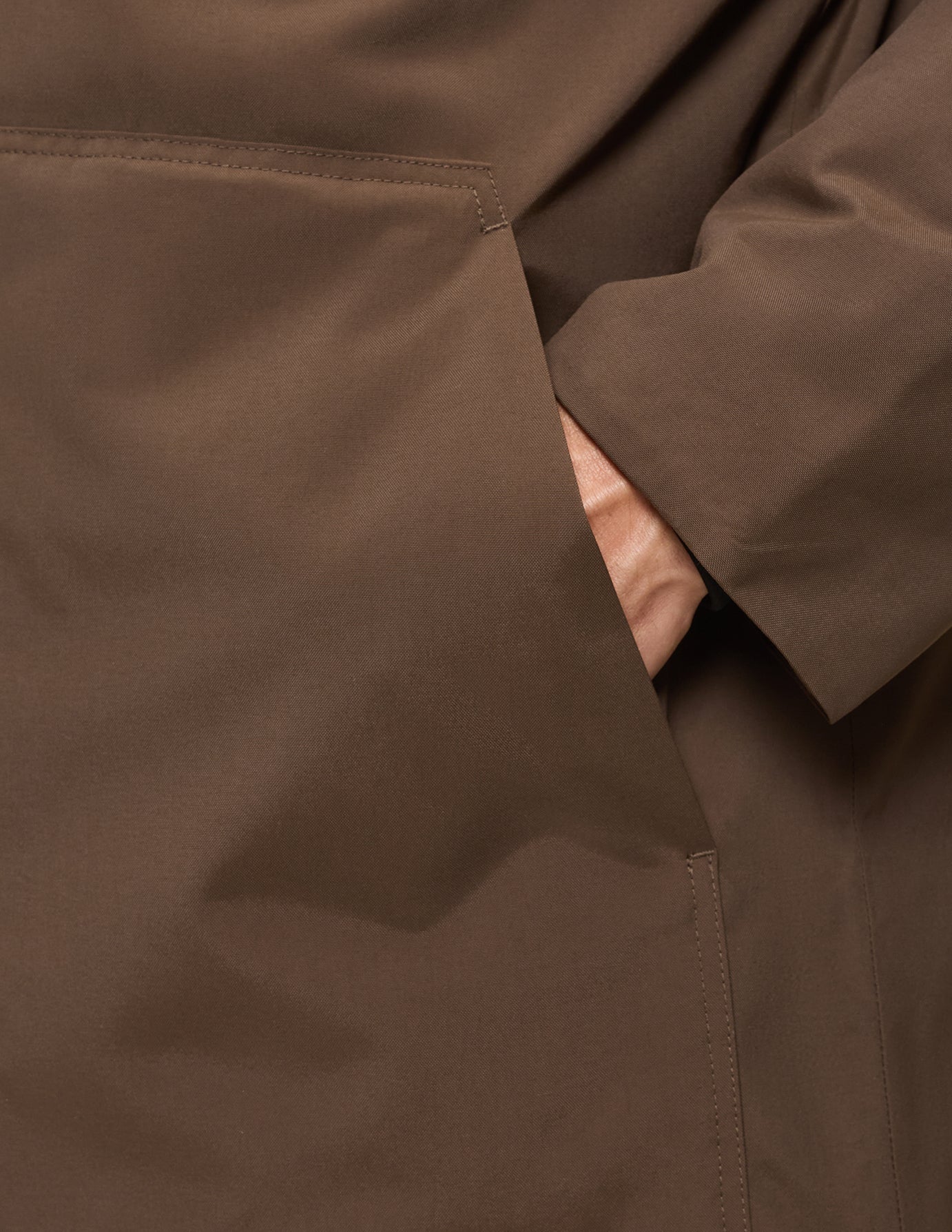 Nærbillede af en hånd, der hviler inde i den store forlomme på en sadelbrun Utility Jacket fremstillet af genanvendt polyester.