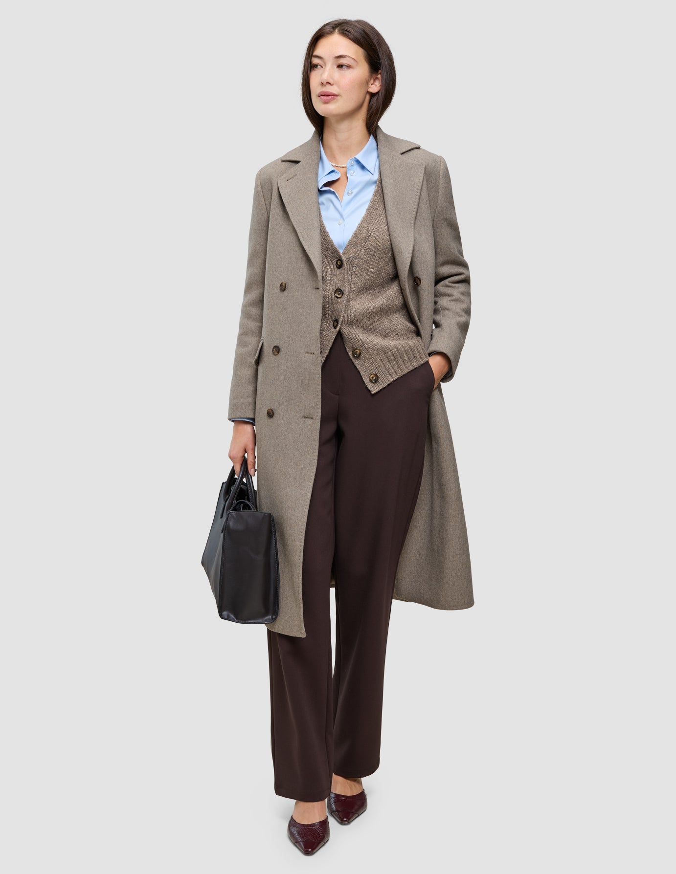 En kvinde iført arbejdstøj til koldt vejr, herunder en beige frakke, en cardigan, en skjorte og bukser, mens hun holder en sort taske.