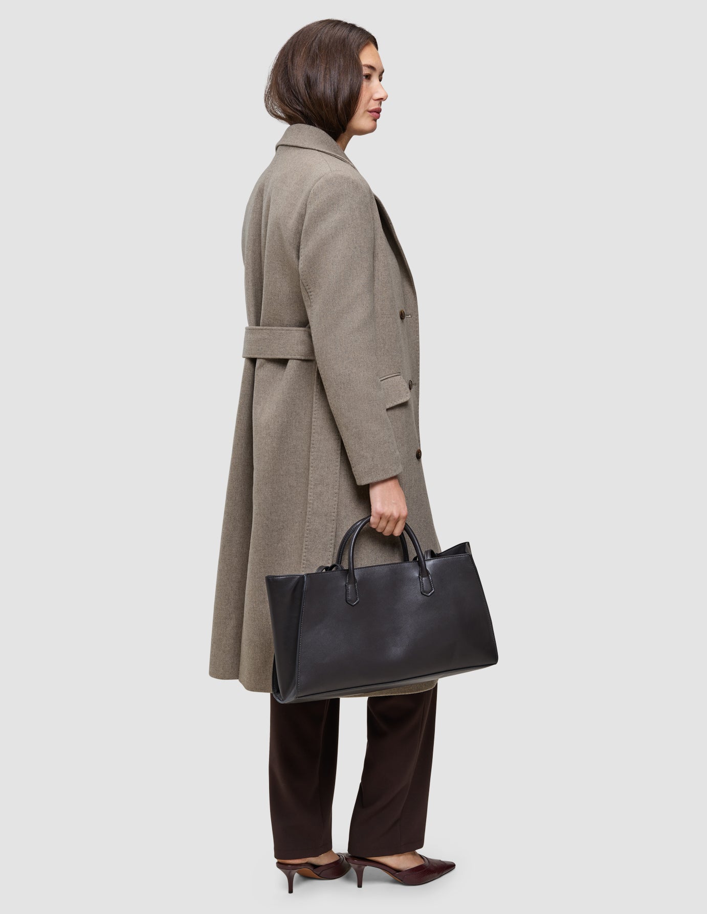 En person, der er iklædt en beige uldfrakke fra serien Cold-weather workwear og mørke bukser, bærer en stor sort totebag.