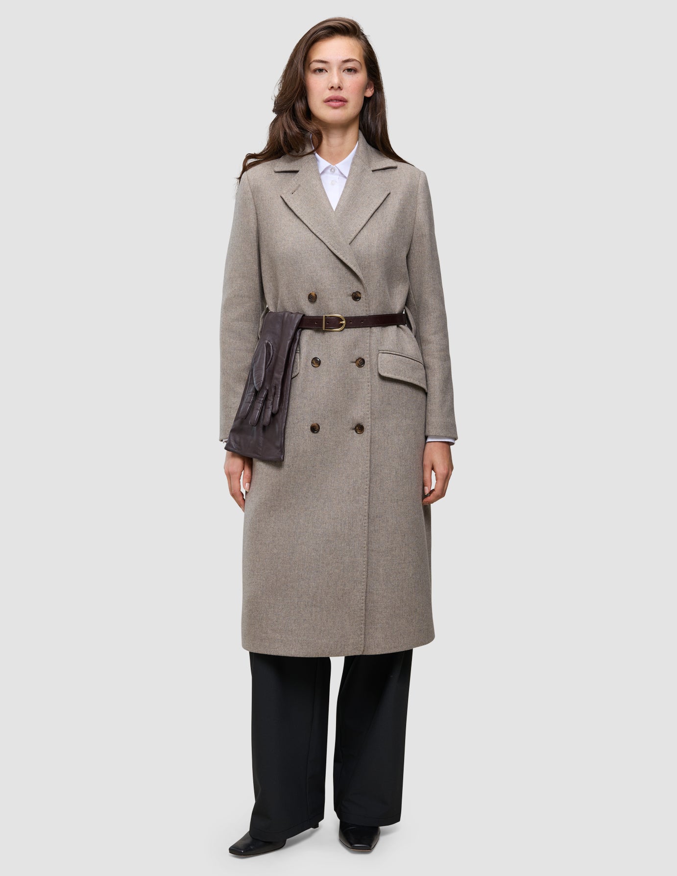 En kvinde bærer en beige uldfrakke med bælte fra Winter, stylet til fest, med mørke handsker stukket ind i taljen for at skabe et klassisk look.