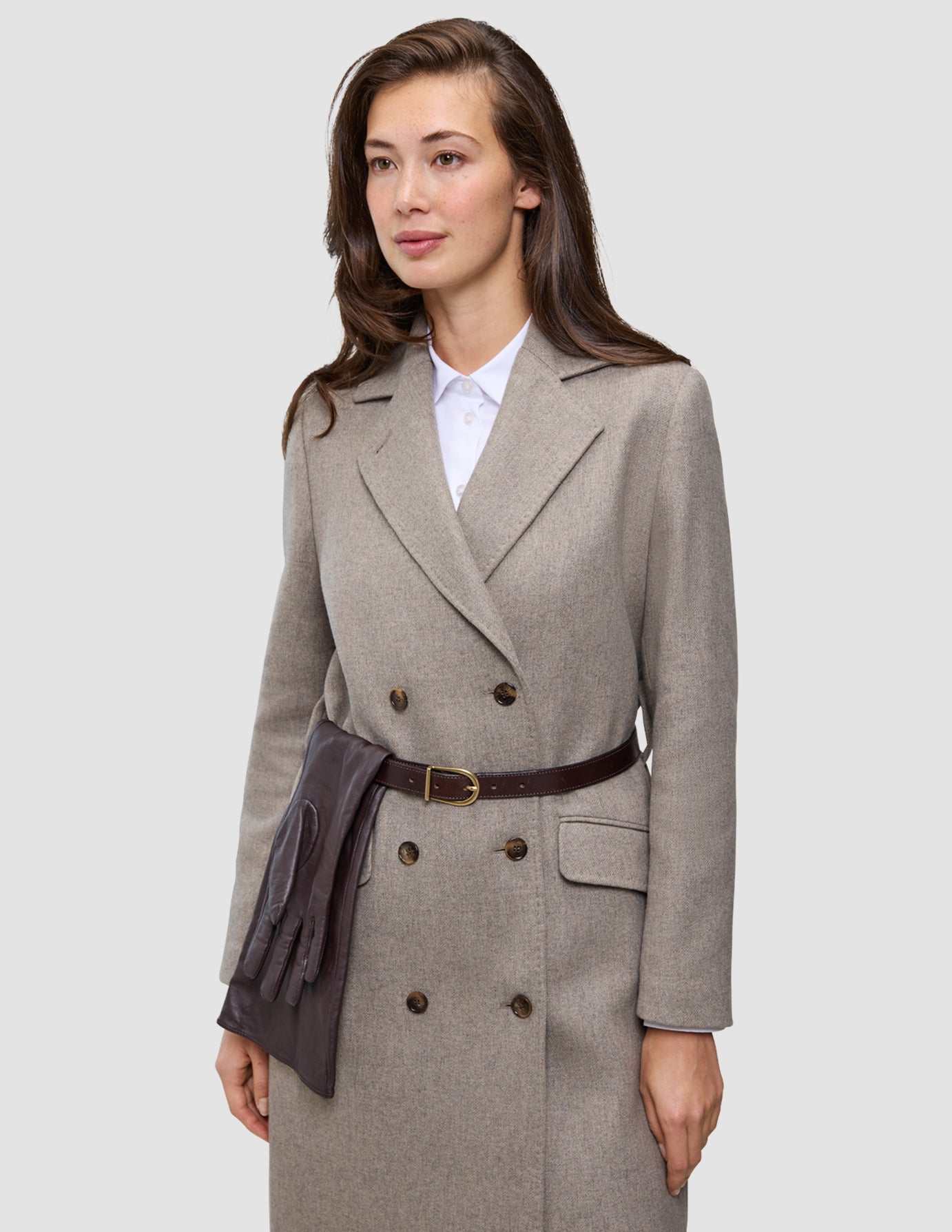 En kvinde har en elegant, beige uldfrakke på over en hvid Oxford-skjorte, og hendes brune læderhandsker er stukket ned i bæltet.