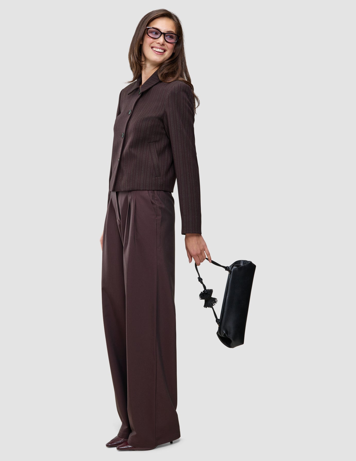 En smilende kvinde er klædt i Burgundy-farven fra top til tå: en mørkebrun Serene Short Jacket med tynde striber og Avira-bukser samt en håndtaske.