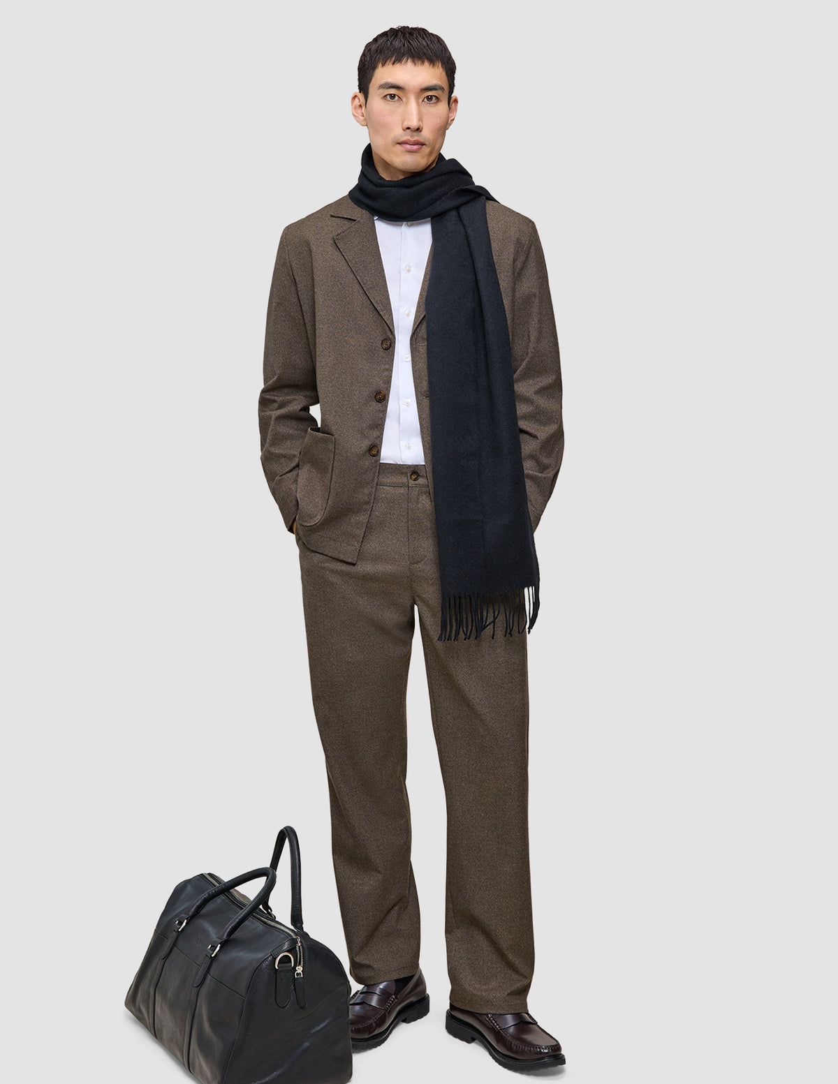 En mand iført en brun Atelier Overshirt Dark Beige, en hvid skjorte og et sort tørklæde står ved siden af en sort læderrejsetaske.